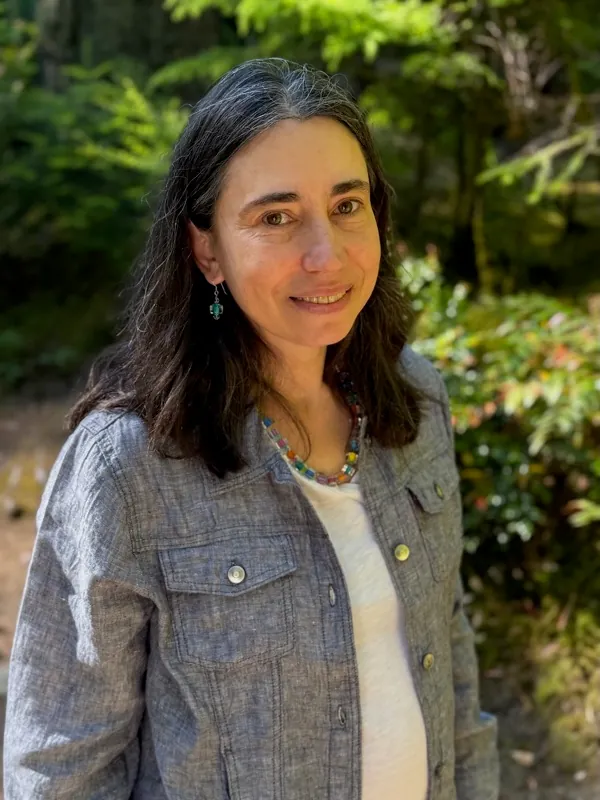 Petia Sierschynski — smiling warmly outdoors, with long dark hair, wearing a chambray jacket over a cream top and a colorful beaded necklace, in front of a lush green forest.